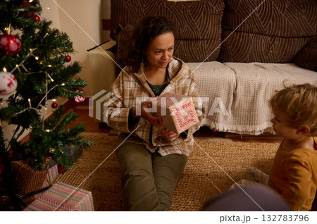 Mother Gifting a Present to Child by the Christmas Tree at Home Mother Gifting a Present to Child by the Christmas Tree at Home 132783796
