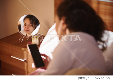 Woman Looks At Her Reflection In A Small Mirror On A Bedroom Dresser While Holding A Phone Woman Looks At Her Reflection In A Small Mirror On A Bedroom Dresser While Holding A Phone 132785299