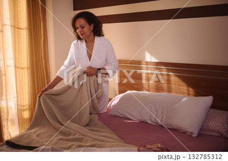 Woman In White Bathrobe Making Bed In Sunny Bedroom With Cozy Blanket 132785312