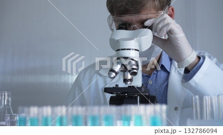 Male scientist, wearing a lab coat, mask, white gloves and safety glasses, is using a binocular microscope surrounded by test tubes in laboratory Male scientist, wearing a lab coat, mask, white gloves and safety glasses, is using a binocular microscope surrounded by test tubes in laboratory 132786094