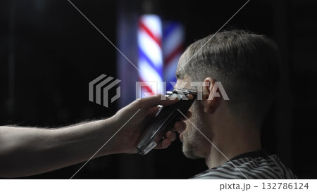 Skilled barber trimming client's hair using electric clipper, vintage barbershop interior with blurred pole, professional grooming service Skilled barber trimming client's hair using electric clipper, vintage barbershop interior with blurred pole, professional grooming service 132786124