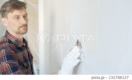 Male construction worker spreading plaster on white wall, wearing protective gloves during home renovation project with putty knife Male construction worker spreading plaster on white wall, wearing protective gloves during home renovation project with putty knife 132786127