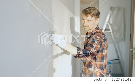 Male construction worker wearing protective gloves smoothing plaster onto wall surface during home renovation using professional trowel tool Male construction worker wearing protective gloves smoothing plaster onto wall surface during home renovation using professional trowel tool 132786148