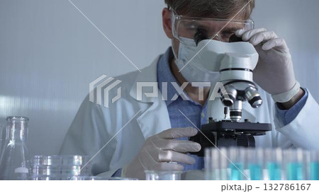 Male scientist, wearing a lab coat, mask, white gloves and safety glasses, is using a binocular microscope surrounded by test tubes in laboratory. Medicine and science concept 132786167