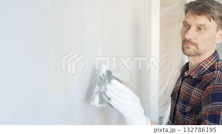 Male construction worker spreading plaster on white wall, wearing protective gloves during home renovation project with putty knife 132786195