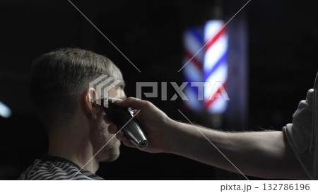 Skilled barber trimming client's hair using electric clipper, vintage barbershop interior with blurred pole, professional grooming service Skilled barber trimming client's hair using electric clipper, vintage barbershop interior with blurred pole, professional grooming service 132786196