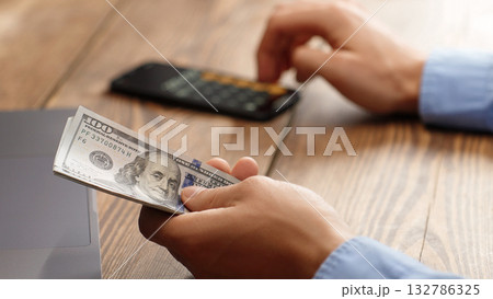 Man holding a pile of 100 dollar bills while using calculator. Financial planning, budgeting or tax calculation concept. Ideal for personal finance, business or investment content. 132786325