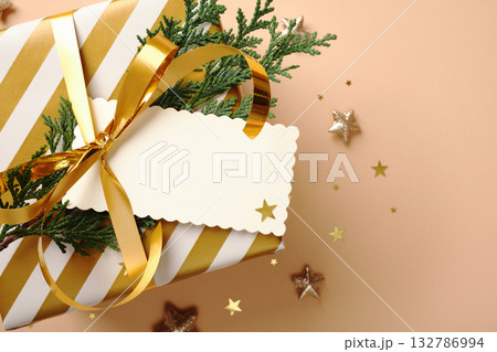 Christmas gift wrapped in gold and white striped paper blank tag, and cedar sprigs on beige background with golden star confetti. Copy space Christmas gift wrapped in gold and white striped paper blank tag, and cedar sprigs on beige background with golden star confetti. Copy space 132786994