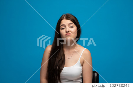 Smug emotion of young woman with raised lip in white tank top on blue background 132788242