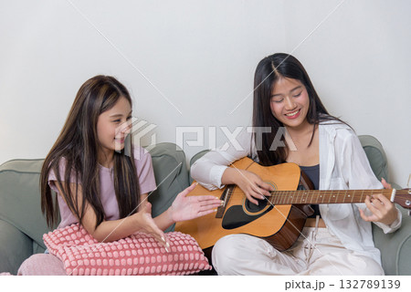 Young girl playing guitar, teen friend attentively watches, women enjoying music session, happy friends bonding, sharing leisure time, creating joyful memories together at home. 132789139