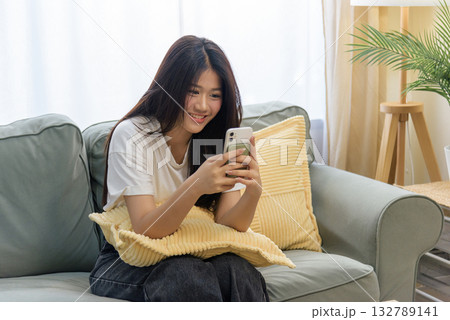 Smiling young Asian woman happy using mobile phone for online communication, person relaxing comfortably on green sofa at home, enjoying cell phone browsing, internet connection, digital lifestyle. 132789141