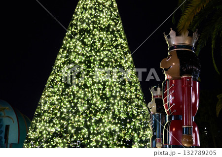 A massive Christmas tree covered in sparkling white and green lights glows in the city at night. Large toy soldier nutcracker statues stand beside the tree while people gather below in winter hats. 132789205