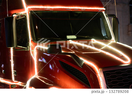 A red tractor trailer cab is traced with glowing white lights along the hood, windshield, and doors at night. The illuminated truck creates a dramatic holiday display on the street. A red tractor trailer cab is traced with glowing white lights along the hood, windshield, and doors at night. The illuminated truck creates a dramatic holiday display on the street. 132789208