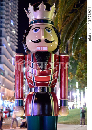A nutcracker soldier statue with a crown, mustache, and red uniform is lit by bright holiday lights in the city at night. The classic Christmas decoration stands in an outdoor public space with people 132789214