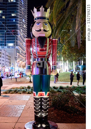 A tall nutcracker soldier statue with a crown stands on a sidewalk in a downtown city at night next to palm trees and holiday lights. People gather in the street behind the festive Christmas A tall nutcracker soldier statue with a crown stands on a sidewalk in a downtown city at night next to palm trees and holiday lights. People gather in the street behind the festive Christmas 132789215
