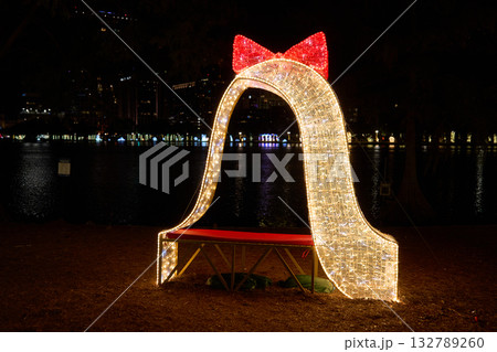 Illuminated arch with a red bow and bench glows beside the city lake at night. Festive holiday decoration with bright LED lights and golden sparkle. 132789260