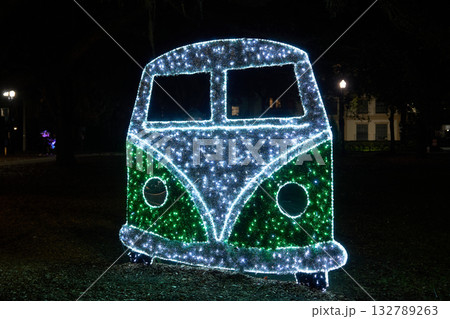 Large illuminated camper van silhouette glows with green and white LED lights in a park. Festive holiday decoration with vintage bus style and sparkling details. 132789263