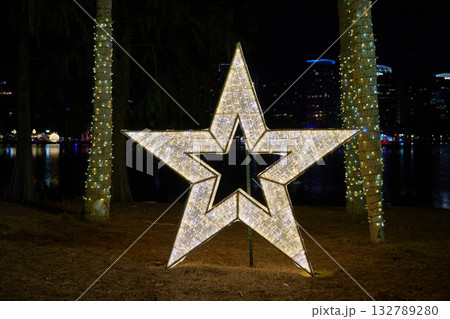 A big glowing star stands between palm trees beside a calm lake at night. Holiday lights, reflections, and skyline create a festive city park scene. 132789280