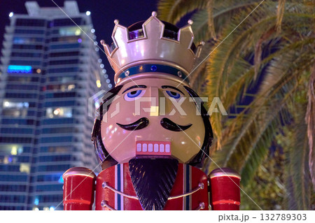 A bright nutcracker figure stands near palm trees and a high-rise building at night. Festive Christmas decoration adds cheerful holiday spirit to the city. 132789303
