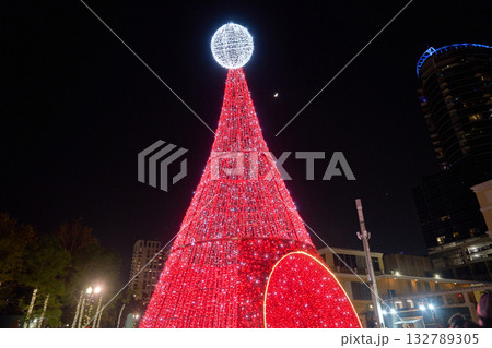 A tall red LED Christmas tree shines brightly against the dark sky in the city. Festive holiday lights create a cheerful downtown winter celebration. A tall red LED Christmas tree shines brightly against the dark sky in the city. Festive holiday lights create a cheerful downtown winter celebration. 132789305