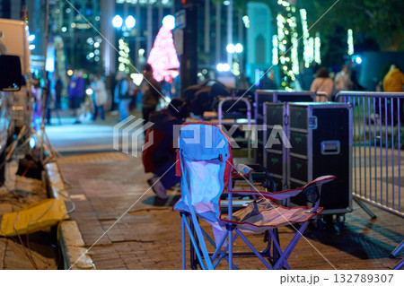A folding chair sits by cases and barricades near news vans on a lit downtown street. The scene shows behind-the-scenes media setup, workflow, and event coverage. A folding chair sits by cases and barricades near news vans on a lit downtown street. The scene shows behind-the-scenes media setup, workflow, and event coverage. 132789307