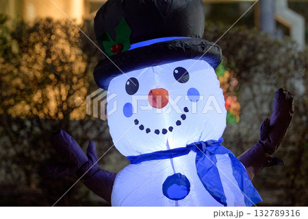 A smiling inflatable snowman wears a black top hat with holly and a blue scarf. The glowing holiday character is captured in a bright night close-up. 132789316