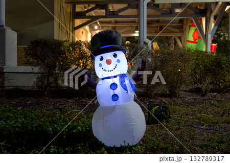 A glowing inflatable snowman with a black top hat and blue scarf stands on the lawn at night. Holiday lights illuminate the cheerful winter decoration in a park setting. A glowing inflatable snowman with a black top hat and blue scarf stands on the lawn at night. Holiday lights illuminate the cheerful winter decoration in a park setting. 132789317