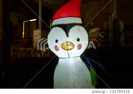 A full inflatable penguin stands outdoors wearing a red Santa hat and scarf. Warm lights illuminate the friendly holiday decoration during a community celebration. 132789318