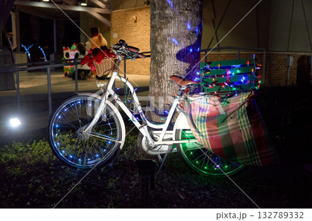 A white city bicycle is decorated with Christmas lights, a poinsettia basket, and a plaid blanket at night. Holiday illumination glows around the wheels and crate near a tree in an outdoor festive 132789332