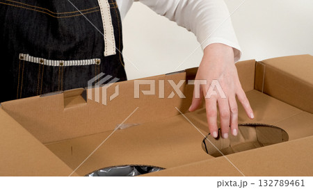Hands unboxing a new cookware set from a cardboard box. White pot with golden handles visible inside the package. 132789461