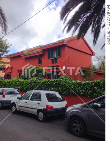 Red house with ivy fence and parked cars in Tenerife. Urban lifestyle, residential architecture, and color harmony of island streets. 132789794