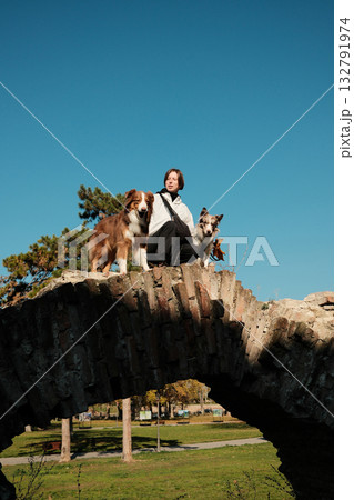 Woman sitting on top of an old stone bridge with two dogs Border Collie and Australian Shepherd under clear blue sky in autumn park Woman sitting on top of an old stone bridge with two dogs Border Collie and Australian Shepherd under clear blue sky in autumn park 132791974