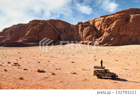 Dusty desert with rocky massif and blue sky above, off road desert vehicle in foreground with young man (unrecognisable, seen from behind) standing on the roof. Typical scenery in Wadi Rum, Jordan 132793221