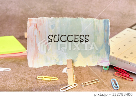 A colorful piece of paper reading SUCCESS is propped up by a clothespin on a textured surface with office supplies such as sticky notes, paperclips, and a calculator nearby A colorful piece of paper reading SUCCESS is propped up by a clothespin on a textured surface with office supplies such as sticky notes, paperclips, and a calculator nearby 132793400