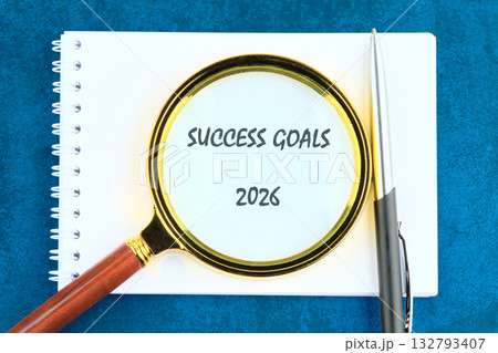 A spiral notebook displays the words Success Goals for 2026, under a gold-rimmed magnifying glass, next to a pen, on a textured blue background 132793407