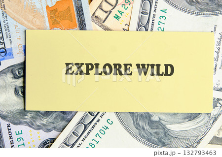 A rectangular piece of light yellow paper with the words EXPLORE WILD printed in dark brown sits atop a bed of United States currency 132793463