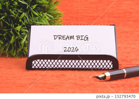 A card that says DREAM BIG 2026 is displayed. Next to the card is a pen, and a green plant is behind it all, on a wooden surface A card that says DREAM BIG 2026 is displayed. Next to the card is a pen, and a green plant is behind it all, on a wooden surface 132793470