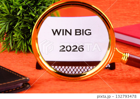 A magnifying glass focuses on a paper that says Win Big 2026. A black notebook and a red book are next to the glass on an orange surface A magnifying glass focuses on a paper that says Win Big 2026. A black notebook and a red book are next to the glass on an orange surface 132793478