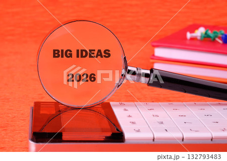 Magnifying glass rests on a calculator, enlarging the words Big Ideas 2026. A stack of red notebooks and push pins sit nearby, all on an orange surface 132793483