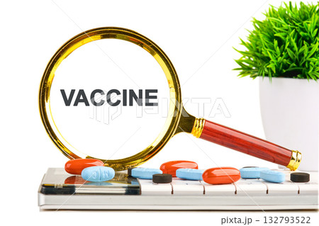 A magnifying glass highlights the word VACCINE on what appears to be a computer screen. Pills are scattered on a keyboard next to a potted plant 132793522