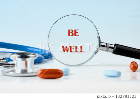 A stethoscope and some pills lay on a white surface. A magnifying glass with the words BE WELL in red letters is centered. Healthcare and wellness are themes 132793523