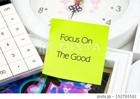 A neon yellow sticky note boldly states Focus On The Good, positioned among a white calculator, a clock face, and picture frames, suggesting a moment of positive reflection 132793582