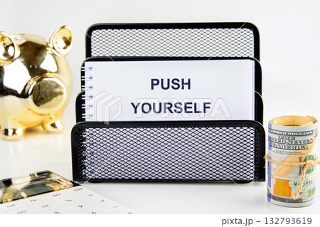 A notebook with the words PUSH YOURSELF sits in a holder, next to a golden piggy bank, a roll of United States currency, and a calculator, suggesting financial motivation 132793619