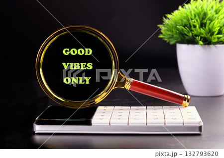 A magnifying glass with GOOD VIBES ONLY on it sits atop a calculator. A small green plant sits in a white pot in the background on a table in front of a black background 132793620