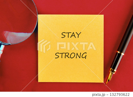 On a red surface, a yellow sticky note displays the phrase Stay Strong. A magnifying glass and a fountain pen rest nearby, suggesting a moment of focus or introspection 132793622