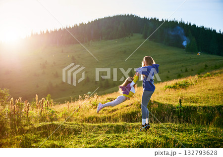 Young woman tourist swings, spending time together with little daughter in sunlit meadow, against stunning mountain backdrop at sunset. Joyful moment captures essence of carefree fun, natural beauty. 132793628
