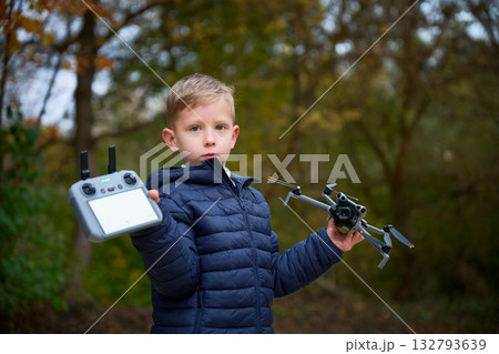 A boy stands in a vibrant autumn setting, confidently holding a drone in one hand and a controller in the other. Leaves in various colors surround him, highlighting the season. A boy stands in a vibrant autumn setting, confidently holding a drone in one hand and a controller in the other. Leaves in various colors surround him, highlighting the season. 132793639