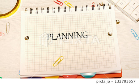 A grid notebook with the word PLANNING on it rests on a table with colorful paperclips, a calculator, and a gold frame. Organization and preparation are implied A grid notebook with the word PLANNING on it rests on a table with colorful paperclips, a calculator, and a gold frame. Organization and preparation are implied 132793657