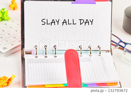 A ring bound planner is open on a desk. The words SLAY ALL DAY are written on a page. Eyeglasses, calculator, and crumpled paper complete the productive scene A ring bound planner is open on a desk. The words SLAY ALL DAY are written on a page. Eyeglasses, calculator, and crumpled paper complete the productive scene 132793672