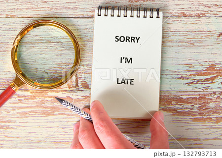A hand holds a notebook open showing the text Sorry Im Late next to a magnifying glass and a patterned pencil on a distressed wooden table A hand holds a notebook open showing the text Sorry Im Late next to a magnifying glass and a patterned pencil on a distressed wooden table 132793713